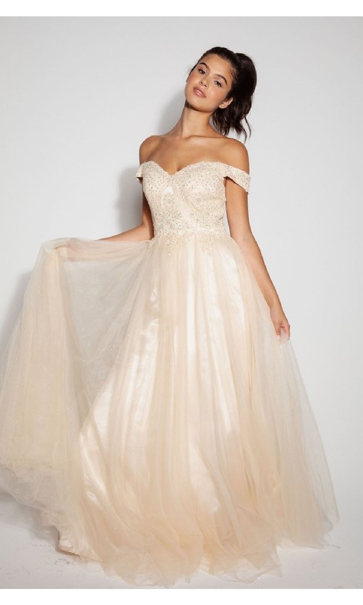 A woman poses in the Eureka 9899 Off-Shoulder Glitter Prom Ball Gown—a floor-length, light beige dress with a fitted embellished bodice and a flowing tulle skirt—against a plain background.